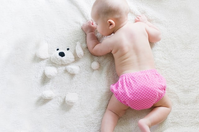 Most Popular Baby Girl Names Right Now, picture of a baby girl laying on her stomach wearing a pink cloth diaper and laying on a white blanket with a stuffed dog attached.